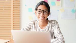 A young person wearing glasses and a striped shirt smiles while sitting at a desk with an open laptop. Colorful sticky notes and papers are visible on the wall behind them.