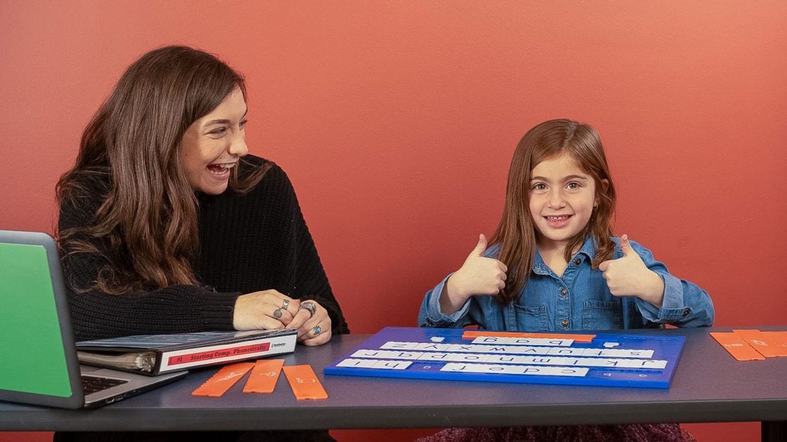 A woman smiles at a young girl who is giving two thumbs up while sitting at a table with letter tiles and educational materials, against a red wall.