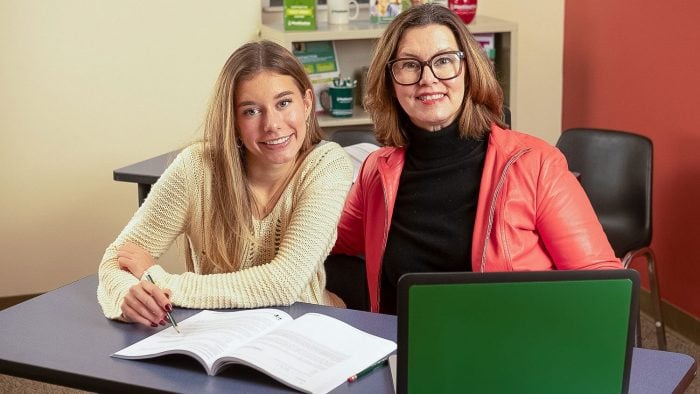 A young student and an older woman sit together at a desk with an open book and a green-laptop. Both are smiling at the camera in a classroom setting.