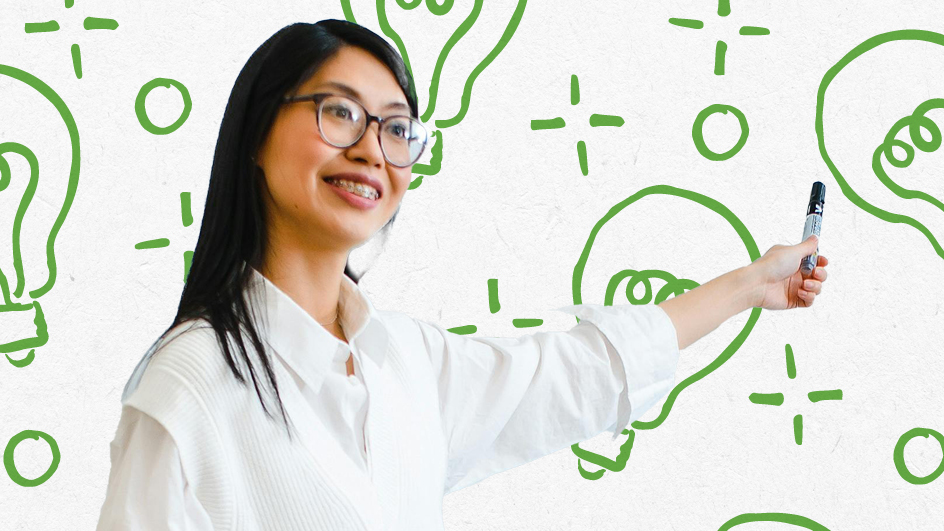 A woman in glasses and a white shirt writes on a whiteboard decorated with large green lightbulb and plus sign illustrations, smiling confidently as she discusses top education trends and holds a marker.