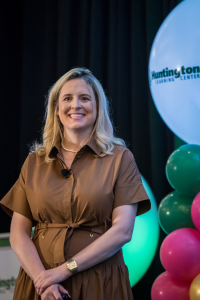 A smiling woman with blonde hair in a brown dress stands before colorful balloons and a Huntington Learning Center sign, speaking to an audience, celebrating as Anne Huntington Sharma is appointed CEO.