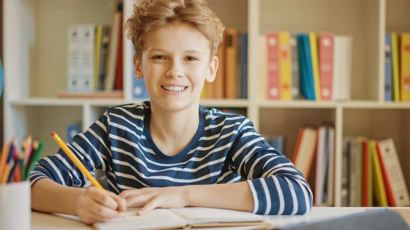 A smiling middle school student wearing a striped shirt sits at a desk, holding a pencil and writing in a notebook, with bookshelves filled with colorful books in the background.