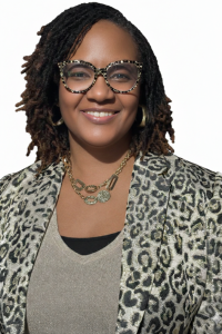 A Huntington franchise owner with shoulder-length, twisted hair wears patterned glasses, gold jewelry, and a leopard-print blazer over a beige top, smiling against a white background.