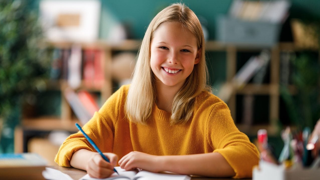 A smiling young girl with blonde hair wearing a yellow sweater sits at a desk, writing in a notebook with a blue pencil. A blurred bookshelf and green wall are visible in the background.