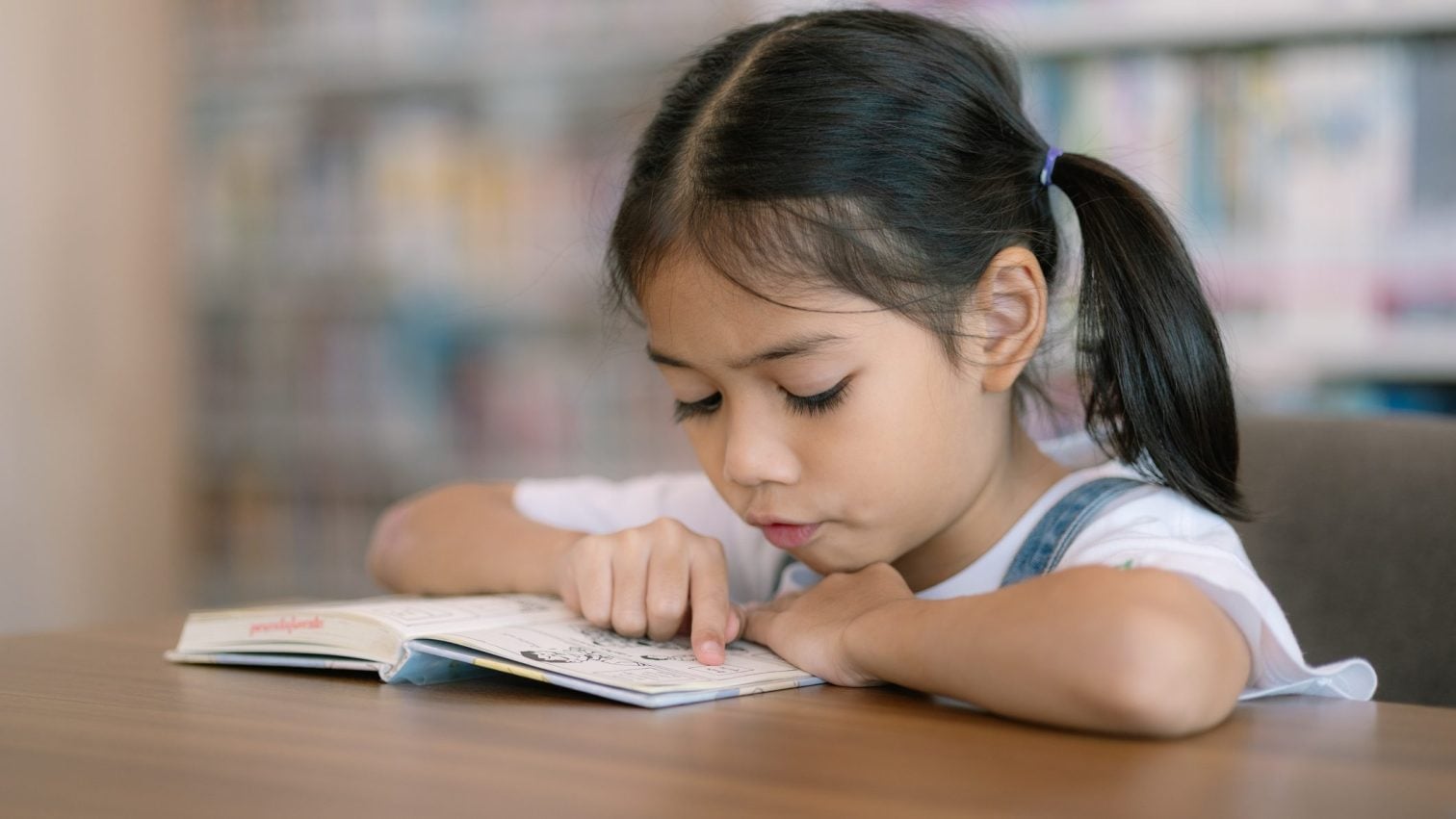 A young girl with dark hair in pigtails sits at a table, intently reading a book—perhaps as part of kindergarten tutoring near me—in a library setting with blurred shelves in the background.