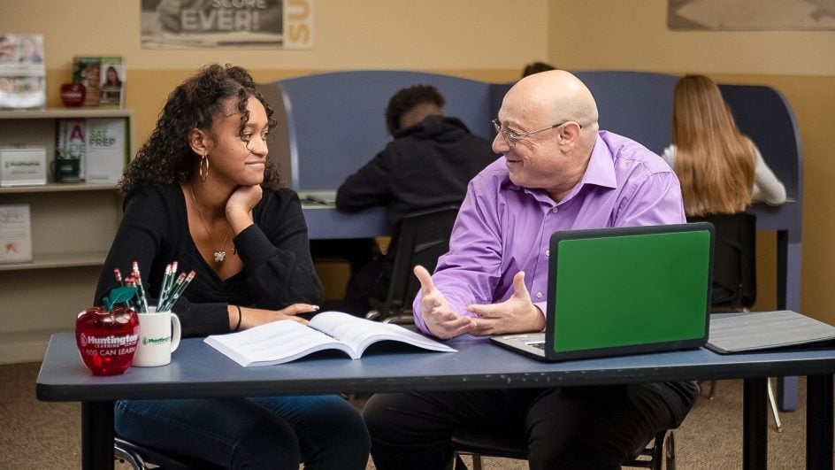 A student and a teacher sit at a table, talking. The student listens with a smile while the teacher gestures. Open books, a laptop, and pens are on the table. Other students work in the background.