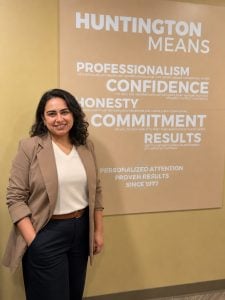 A woman with dark curly hair, wearing a tan blazer and white top, stands smiling in front of a beige wall with motivational words like Professionalism, Confidence, Honesty, and Commitment displayed on it.