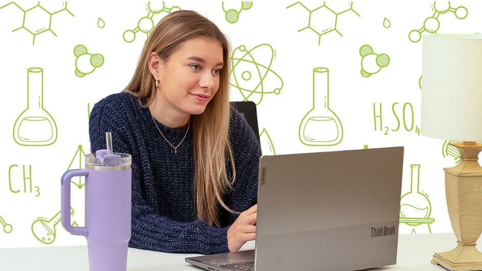 A woman with long blonde hair works on a laptop at a desk. A large purple tumbler and a lamp sit nearby. The background features green chemistry-themed doodles, including beakers, molecules, and atomic symbols.