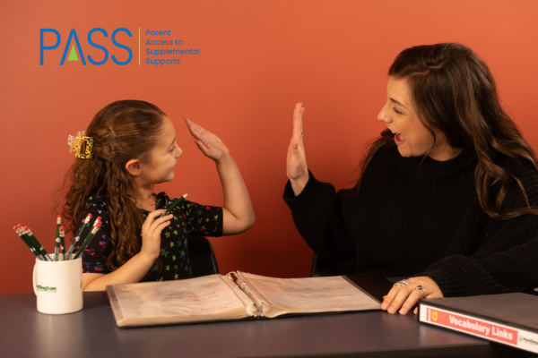 A young girl and a woman smile and give each other a high five across a desk with an open binder, pencils in a cup, and a vocabulary book. The PASS program logo is visible on the wall behind them, highlighting their participation in the Texas Education Freedom Account initiative.